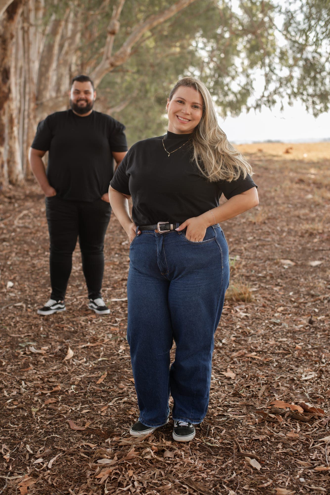 Nâmora sorrindo em primeiro plano e Lucas ao fundo, entre os eucaliptos
